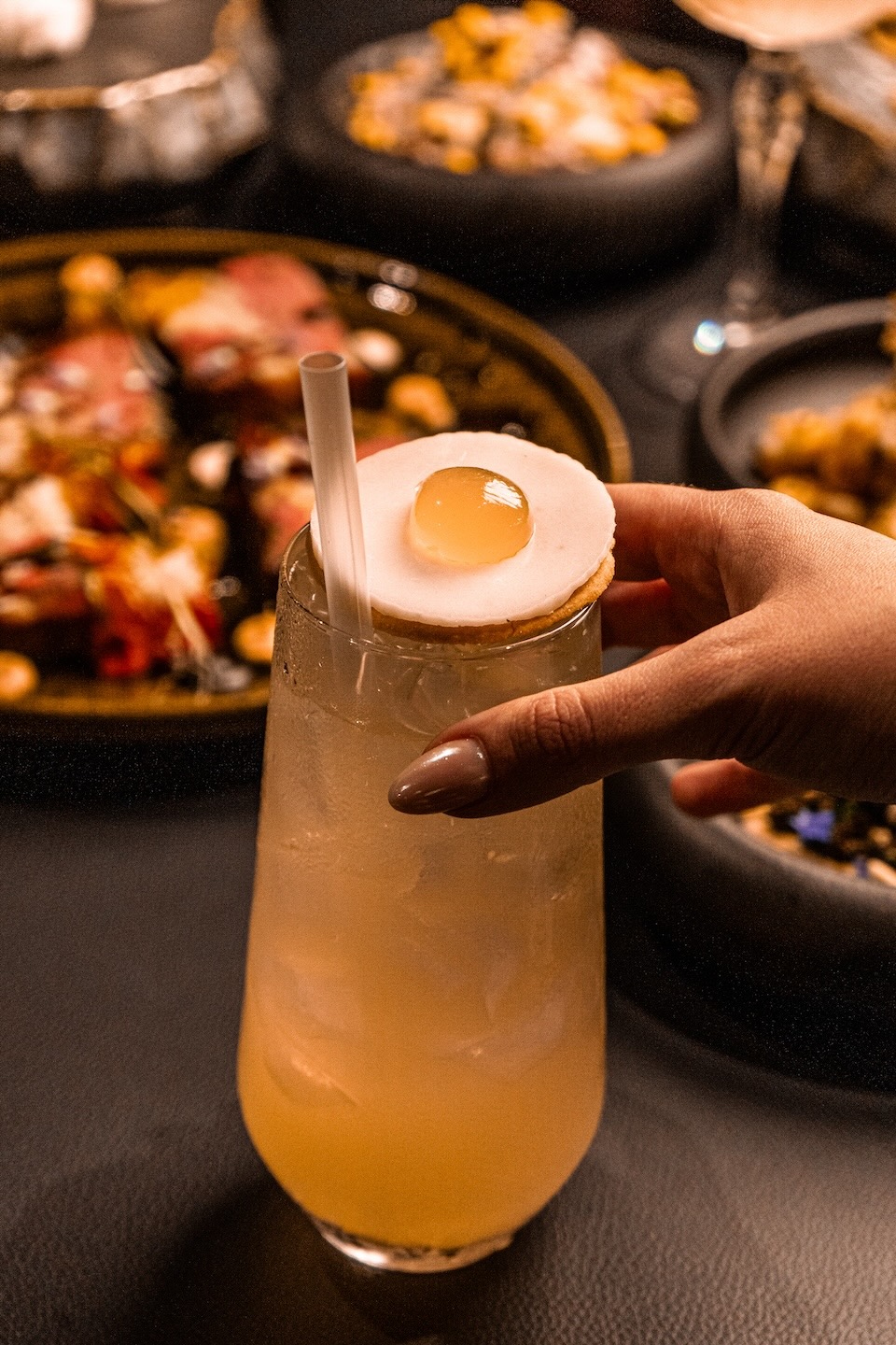 A tall, frosted glass containing a pale yellow cocktail with a white straw. A hand is placing a unique garnish on top that looks like a miniature fried egg sitting on a cracker.