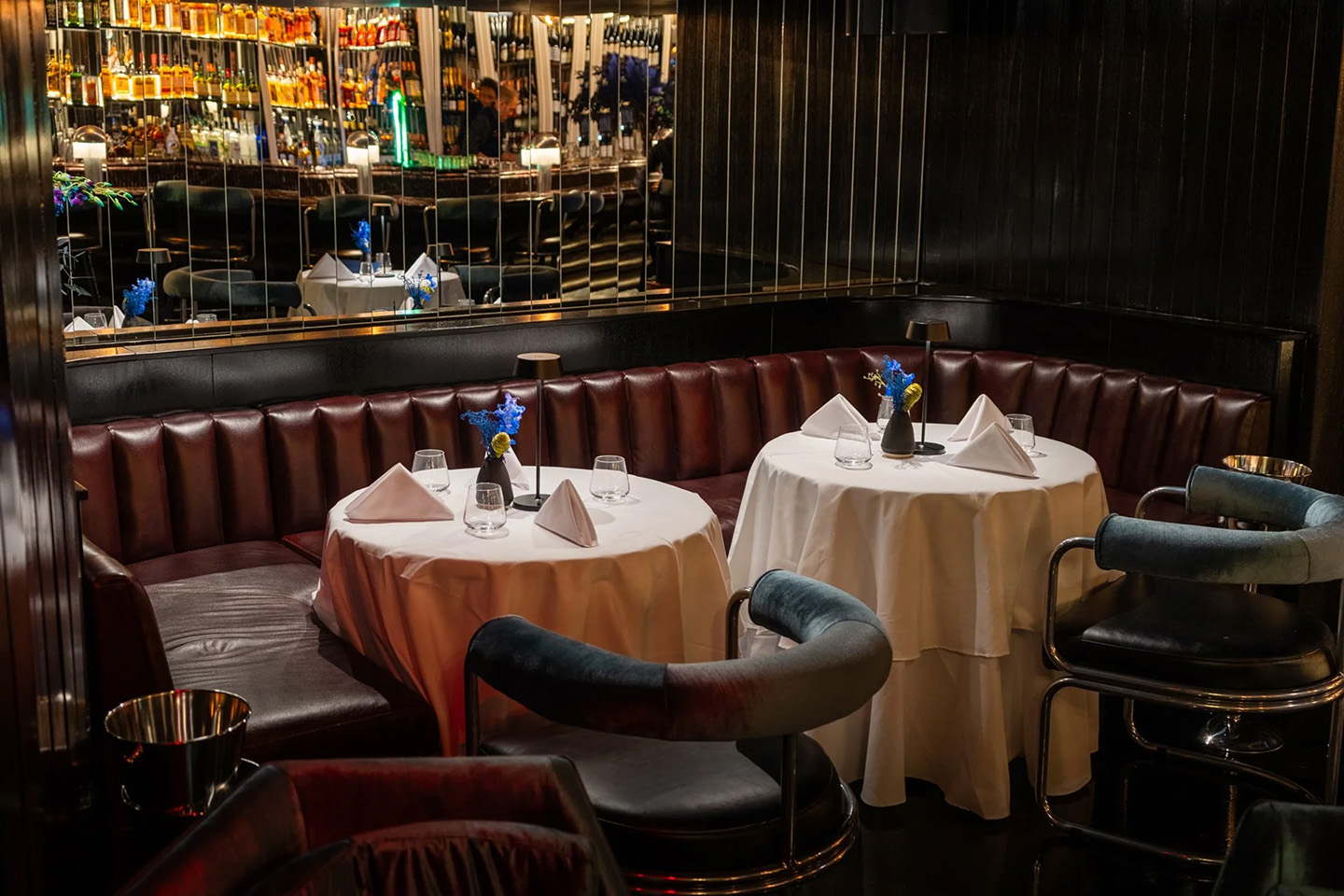 A wide view of a stylish lounge interior featuring dark polished floors, teal velvet chairs, and a long curved leather banquette.