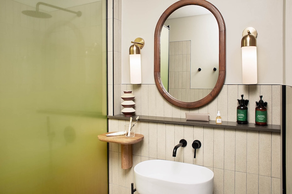 A modern bathroom vanity featuring a white vessel sink with matte black wall-mounted faucets. Above the sink is a large oval mirror with a dark wood frame, flanked by two brass and frosted glass sconces. The wall is finished with vertical cream-colored tiles and a dark stone ledge holding luxury toiletries.