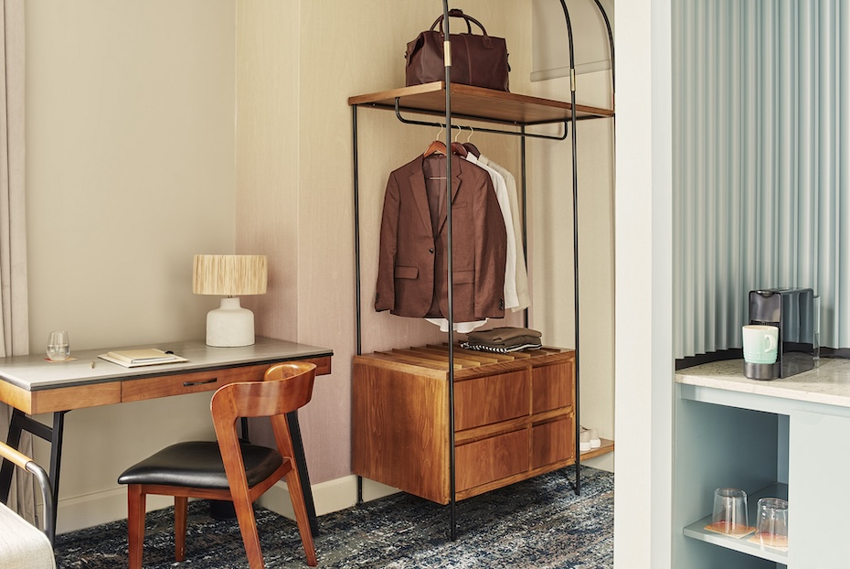 A hotel room workspace and storage area. It features a mid-century modern wooden desk with a black leather chair and a minimalist open-frame wardrobe in black metal and wood. To the right, a partial view of a blue coffee station with a capsule machine and glassware is visible.