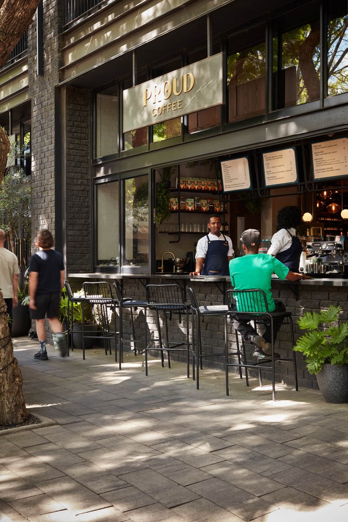 An outdoor view of "Proud Coffee" featuring a black brick walk-up counter with barstools. Staff in aprons work behind the counter while customers walk along the tree-lined sidewalk.