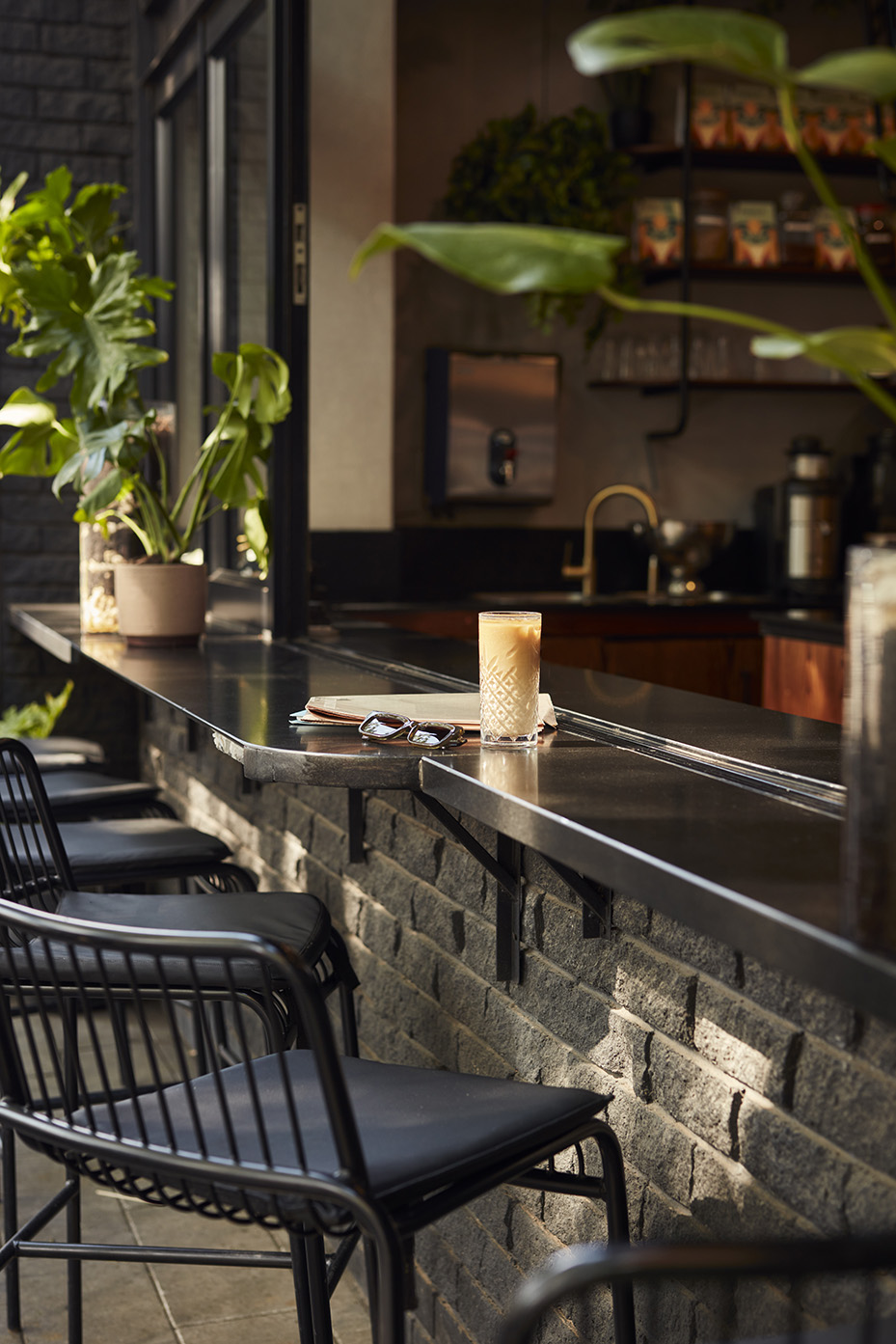 A close-up of a sleek black outdoor counter at a café, featuring an iced coffee in a tall glass, a pair of sunglasses, and a newspaper. Modern black metal barstools and lush green plants line the brick wall.