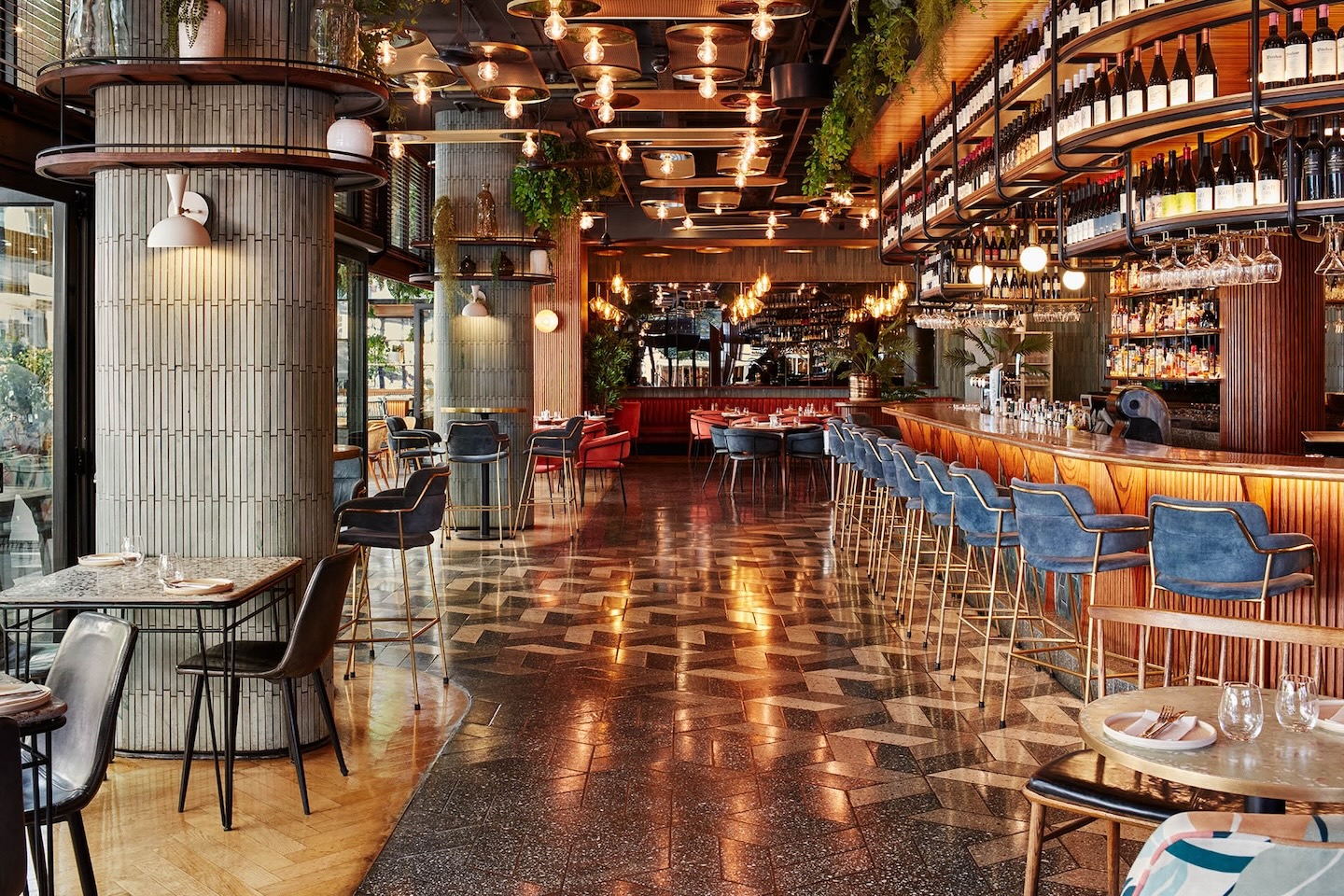 A perspective shot looking down the length of the restaurant, highlighting the contrast between the dark geometric floor tiles and the light wood chevron flooring. Large, cylindrical pillars clad in light grey tiles act as room dividers beneath a ceiling of warm, industrial-style lighting.