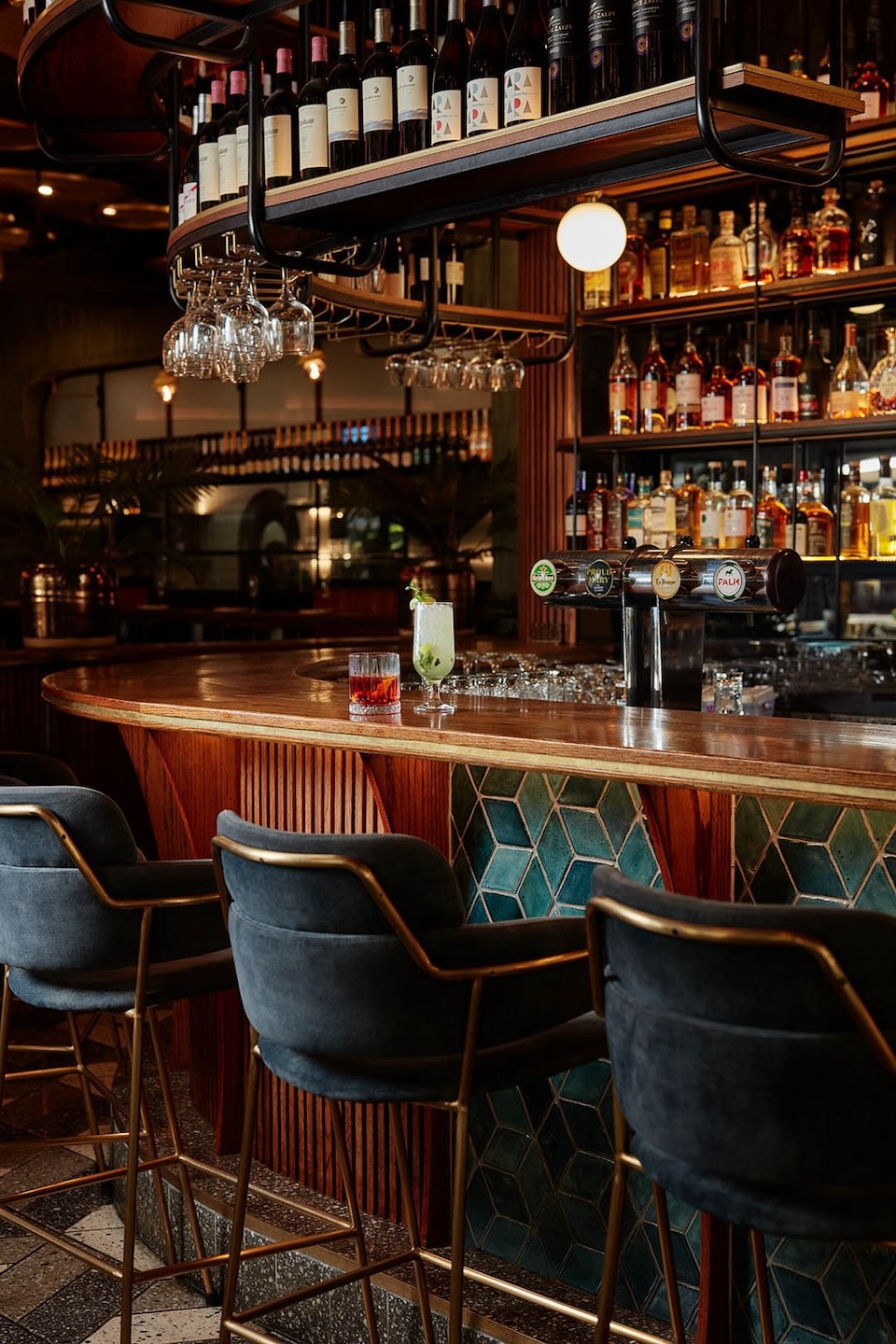 A curved wooden bar featuring a base of teal 3D diamond-shaped tiles and vertical wood slats. Plush charcoal velvet bar stools with gold legs are lined up. On the counter sit a red cocktail and a tall green garnish drink next to beer taps.