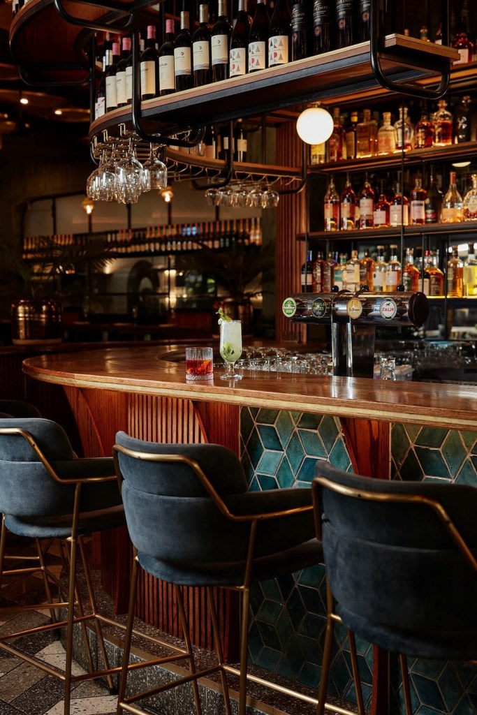 A curved wooden bar featuring a base of teal 3D diamond-shaped tiles and vertical wood slats. Plush charcoal velvet bar stools with gold legs are lined up. On the counter sit a red cocktail and a tall green garnish drink next to beer taps.