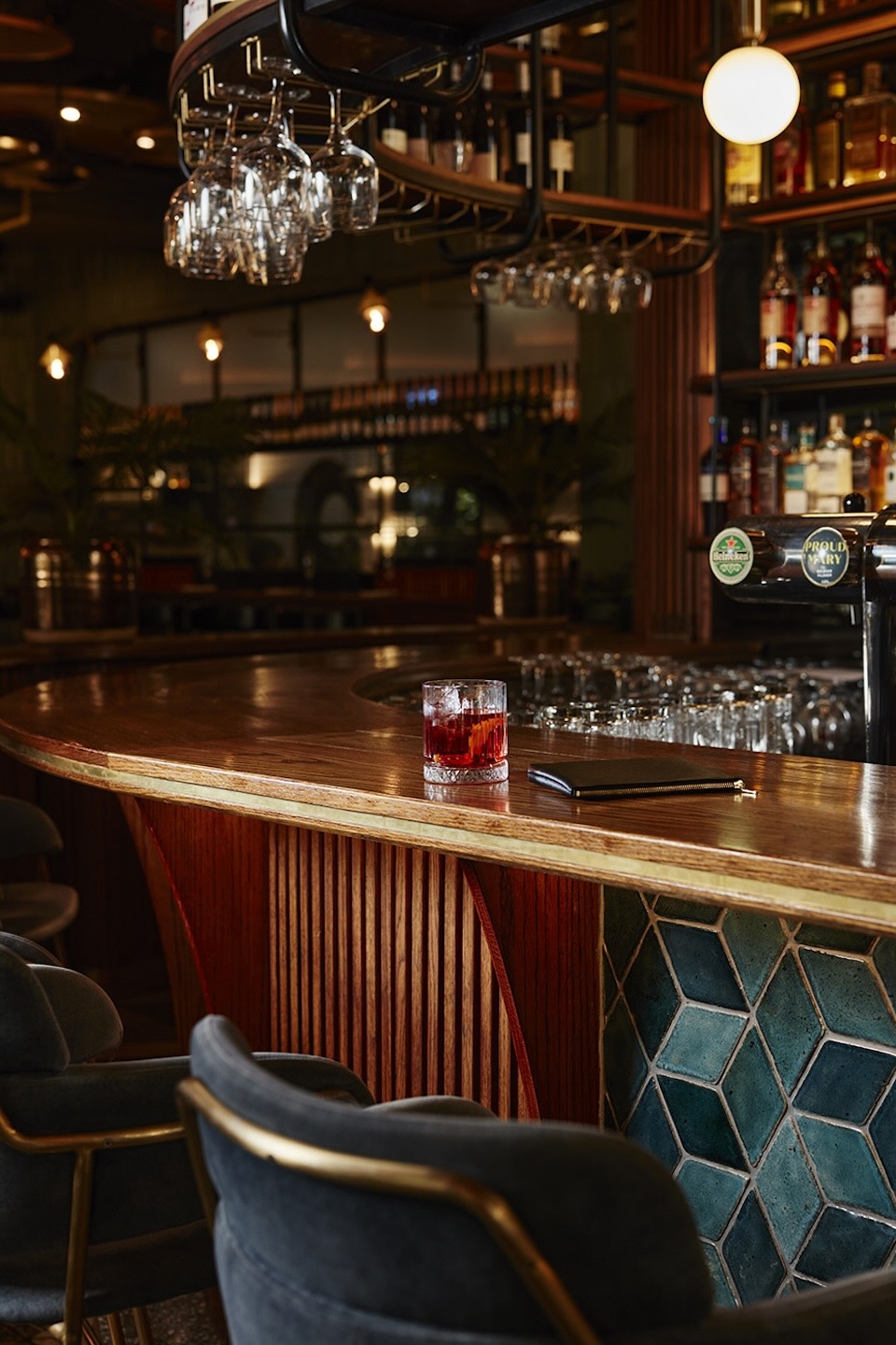A detailed shot of the curved bar counter showing a red cocktail in a crystal glass and a black leather wallet. In the background, rows of wine glasses hang beneath a shelf of spirits, illuminated by warm, spherical pendant lights.