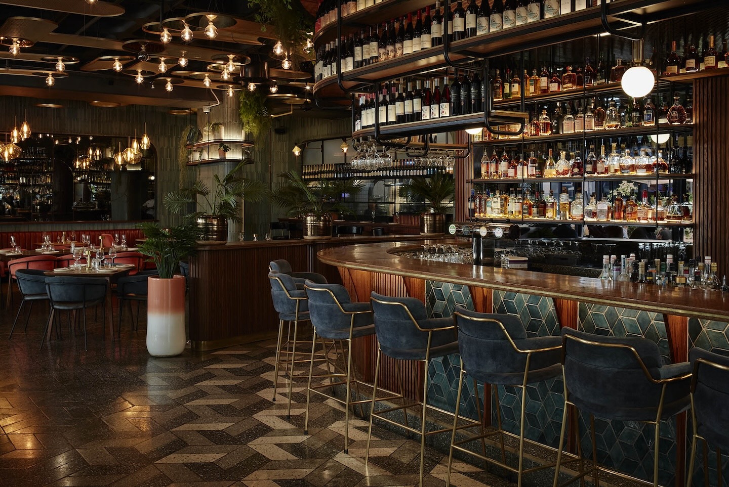 A wide view of the bar area showcasing the extensive backlit shelving filled with liquor and wine. The space features mid-century modern accents, including a patterned terrazzo floor, greenery in large planters, and a glimpse of red velvet dining booths.