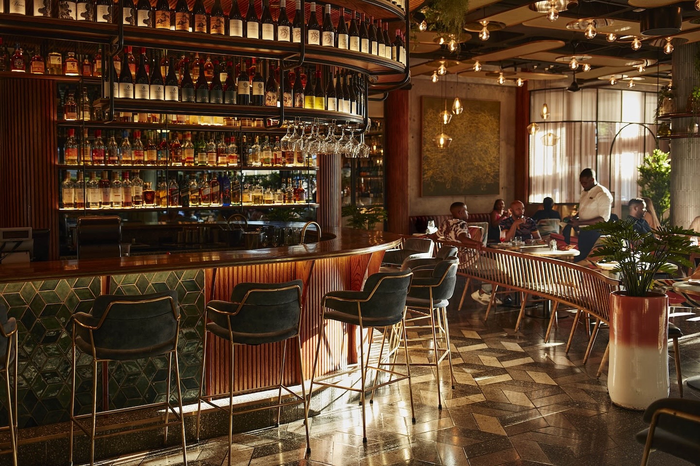 A perspective shot behind the curved bar, highlighting the warm glow of the backlighting on the spirit collection. The foreground features velvet bar stools, while the background shows a dining area with wooden slat benches and patrons enjoying a meal.