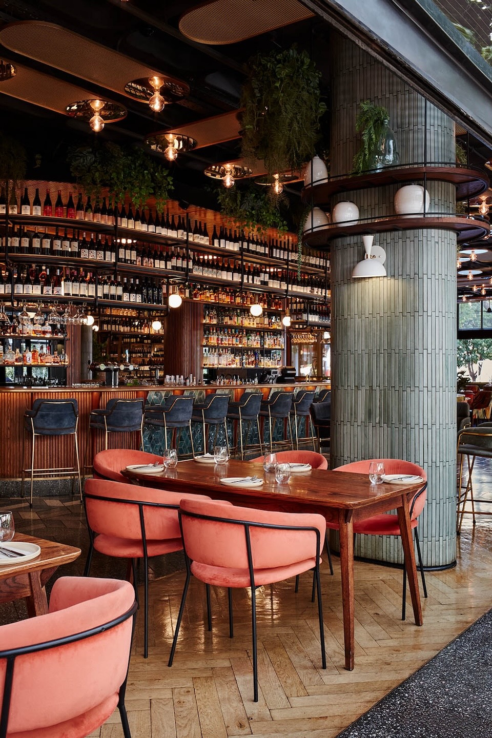 A view of a wooden dining table surrounded by salmon-colored velvet chairs with black metal frames. A large, grey tiled pillar with built-in shelving and plants anchors the room. The expansive bar and wine display stretch across the background.