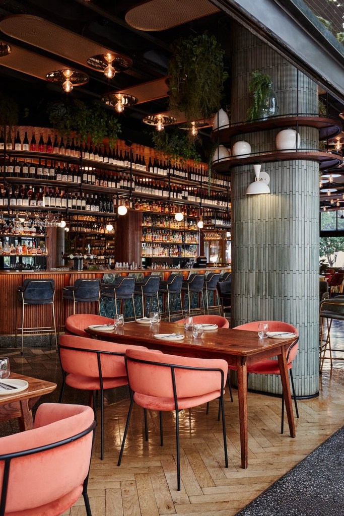 A view of a wooden dining table surrounded by salmon-colored velvet chairs with black metal frames. A large, grey tiled pillar with built-in shelving and plants anchors the room. The expansive bar and wine display stretch across the background.