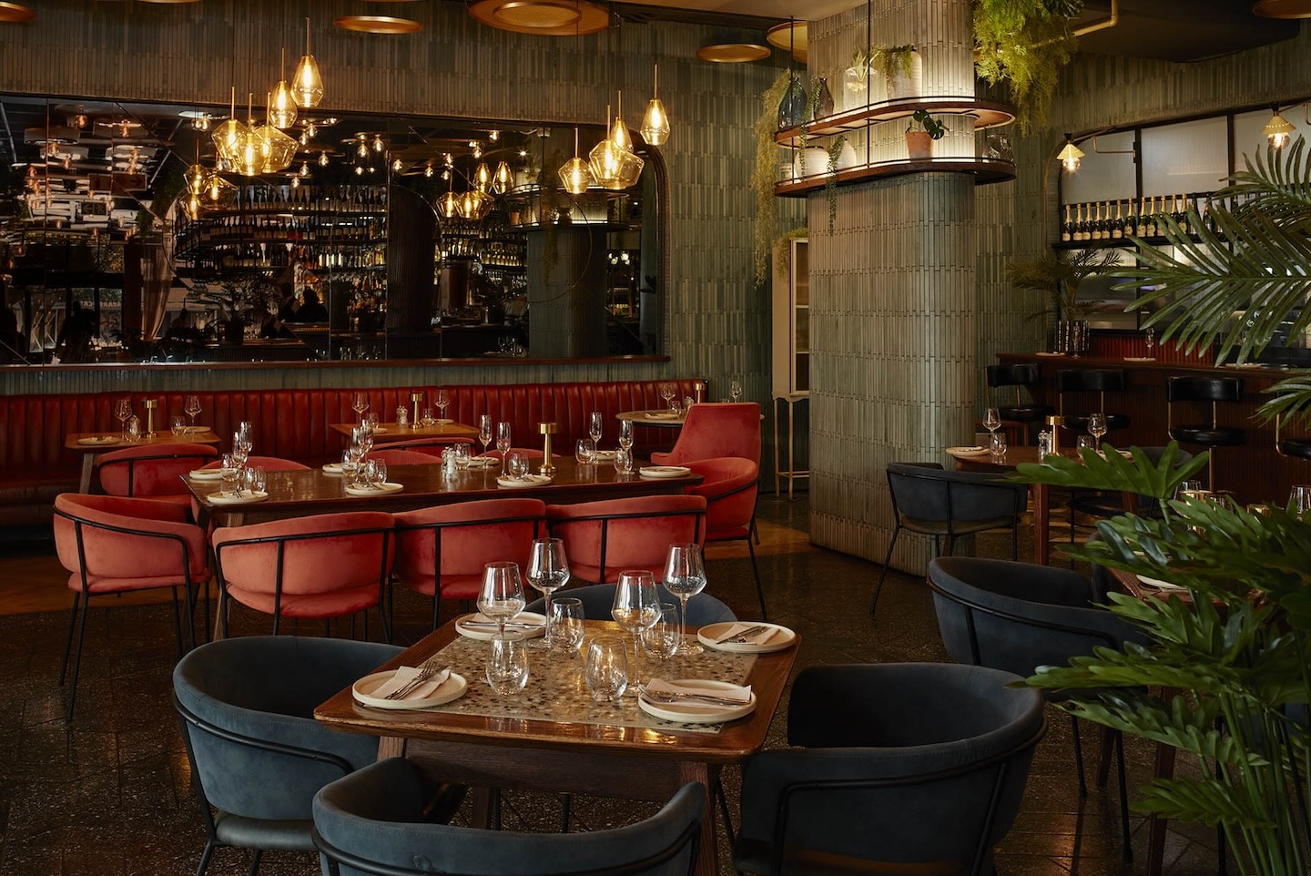 A cozy dining nook featuring a long, red leather tufted banquet against a wall of vertical grey tiles. Groupings of wooden tables and velvet chairs sit under amber-toned glass pendant lights. A large mirror reflects the warm lighting of the restaurant.