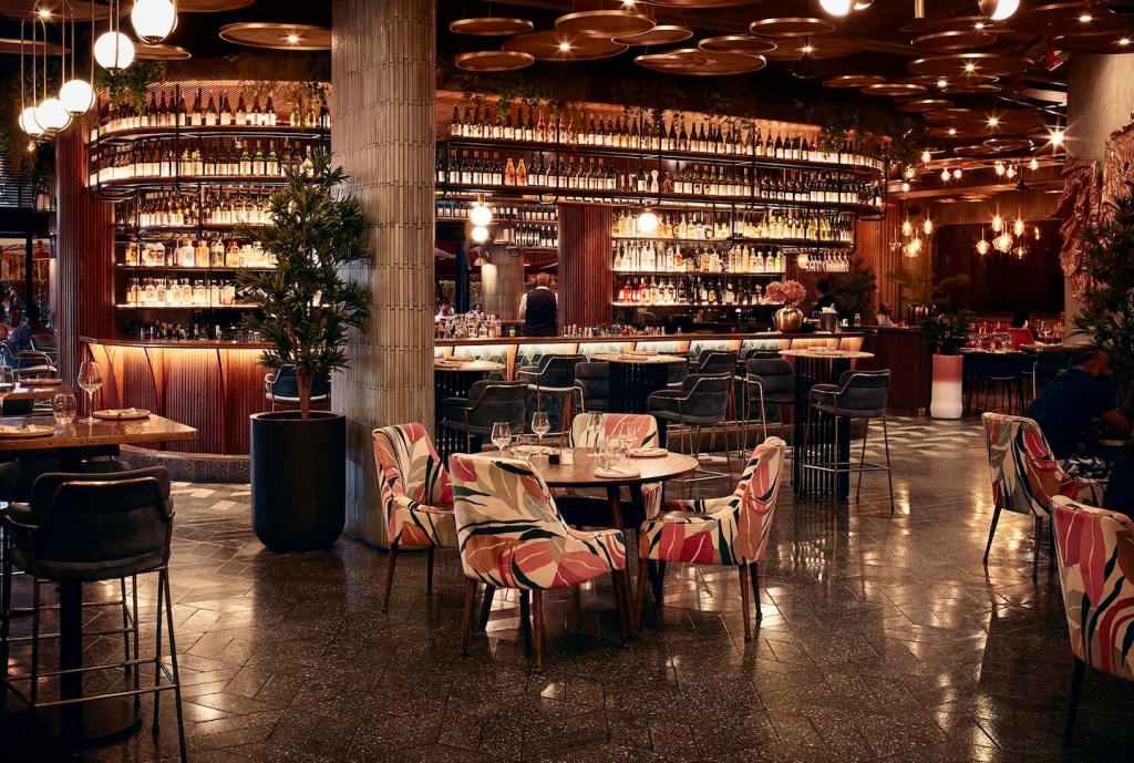 An expansive view of the main floor showing various seating options, including chairs with a bold pink and leaf-patterned print. The ceiling is decorated with unique circular wooden panels and hanging bulbs, creating a layered, textured lighting effect.