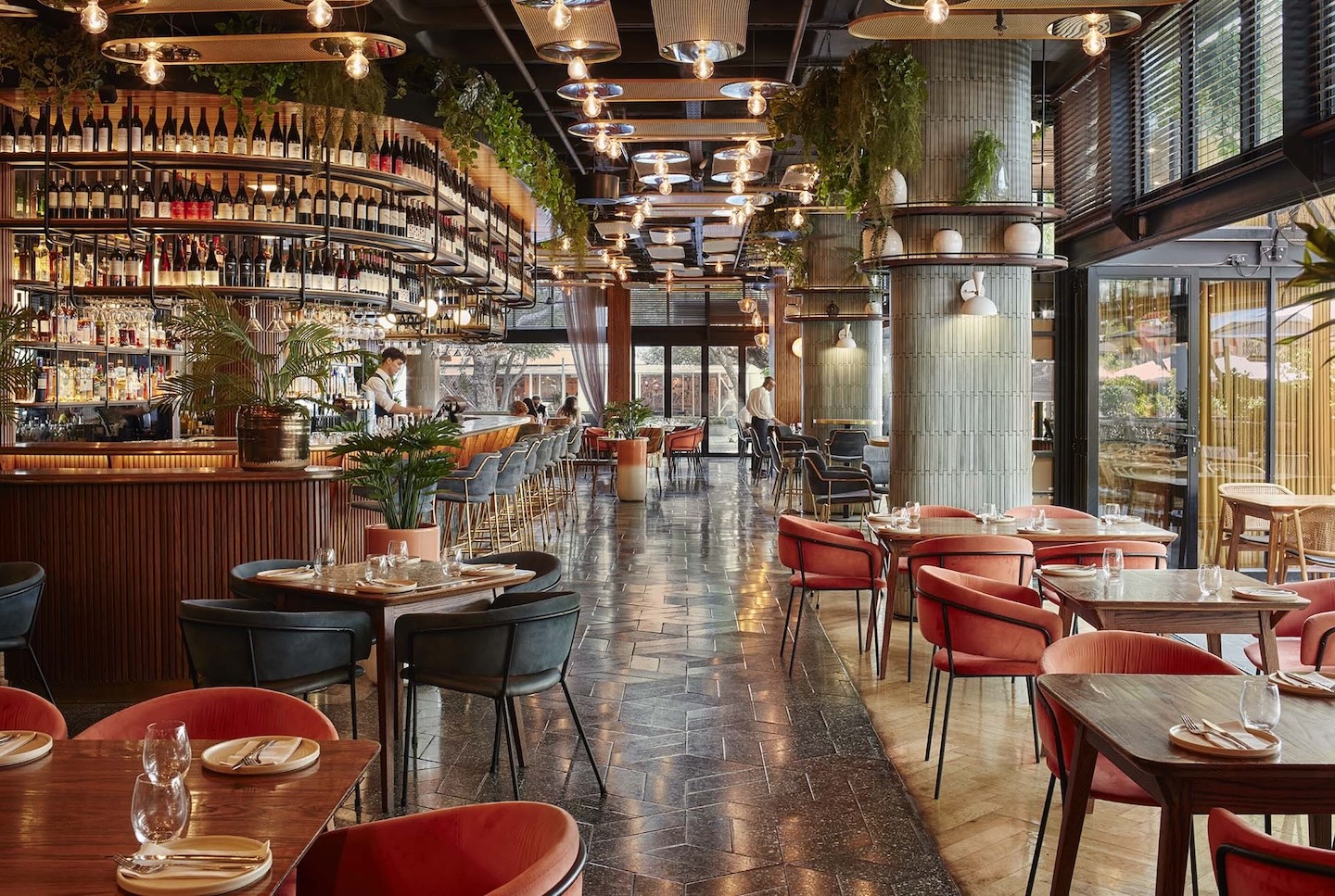 A wide-angle view of the bustling restaurant interior featuring a long, high-top bar with teal velvet stools. Salmon-colored velvet chairs surround dark wood tables in the foreground, while floor-to-ceiling windows and lush hanging plants create an airy, indoor-outdoor feel.