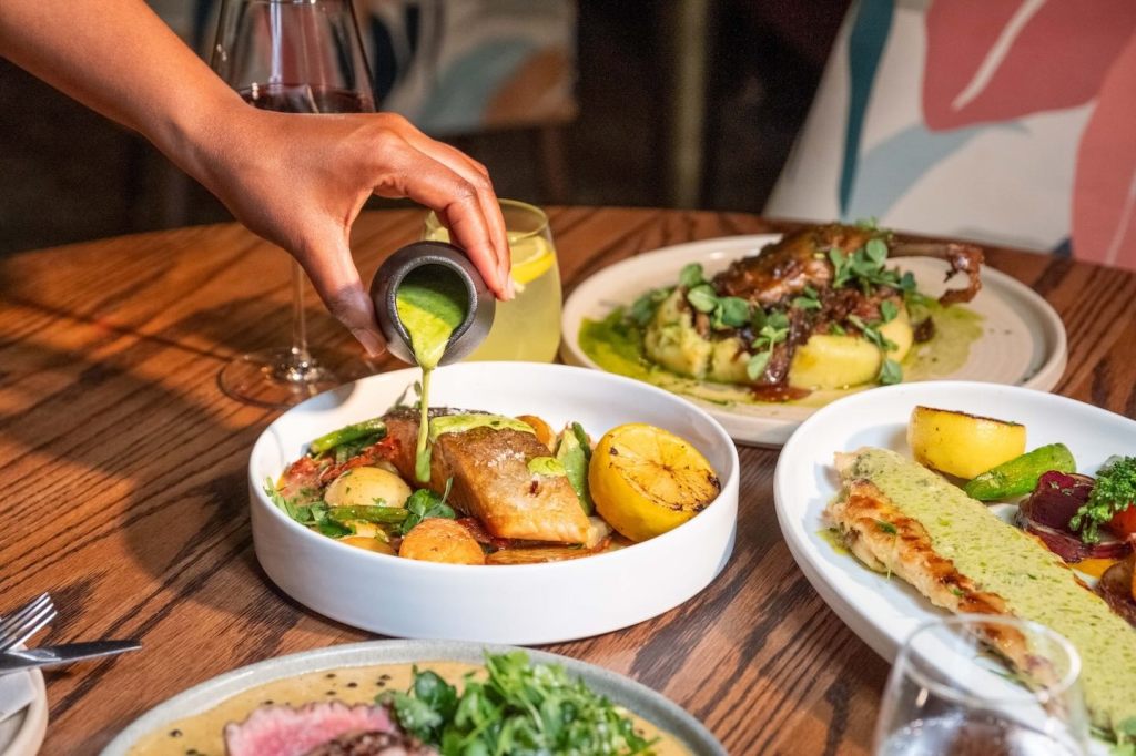 A close-up shot of a hand pouring a vibrant green herb sauce from a small black pitcher onto a pan-seared salmon fillet. The salmon is served in a white bowl with roasted baby potatoes and green beans.