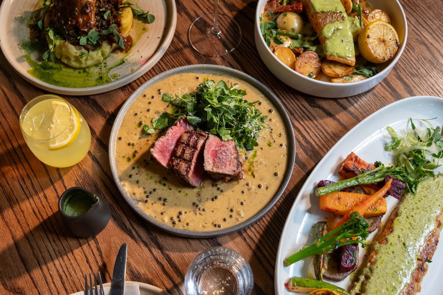 A high-angle view of a wooden dining table showcasing a medium-rare sliced steak served in a peppercorn cream sauce. Surrounding dishes include roasted root vegetables, seared salmon, and a duck confit dish.
