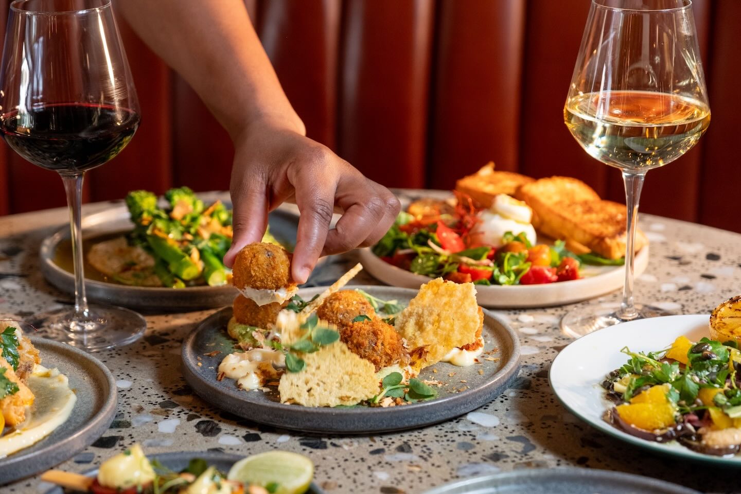 A close-up of a hand picking up a golden-brown arancini ball from a grey plate. The dish includes several crispy croquettes served with creamy aioli and thin, crispy parmesan tuiles.
