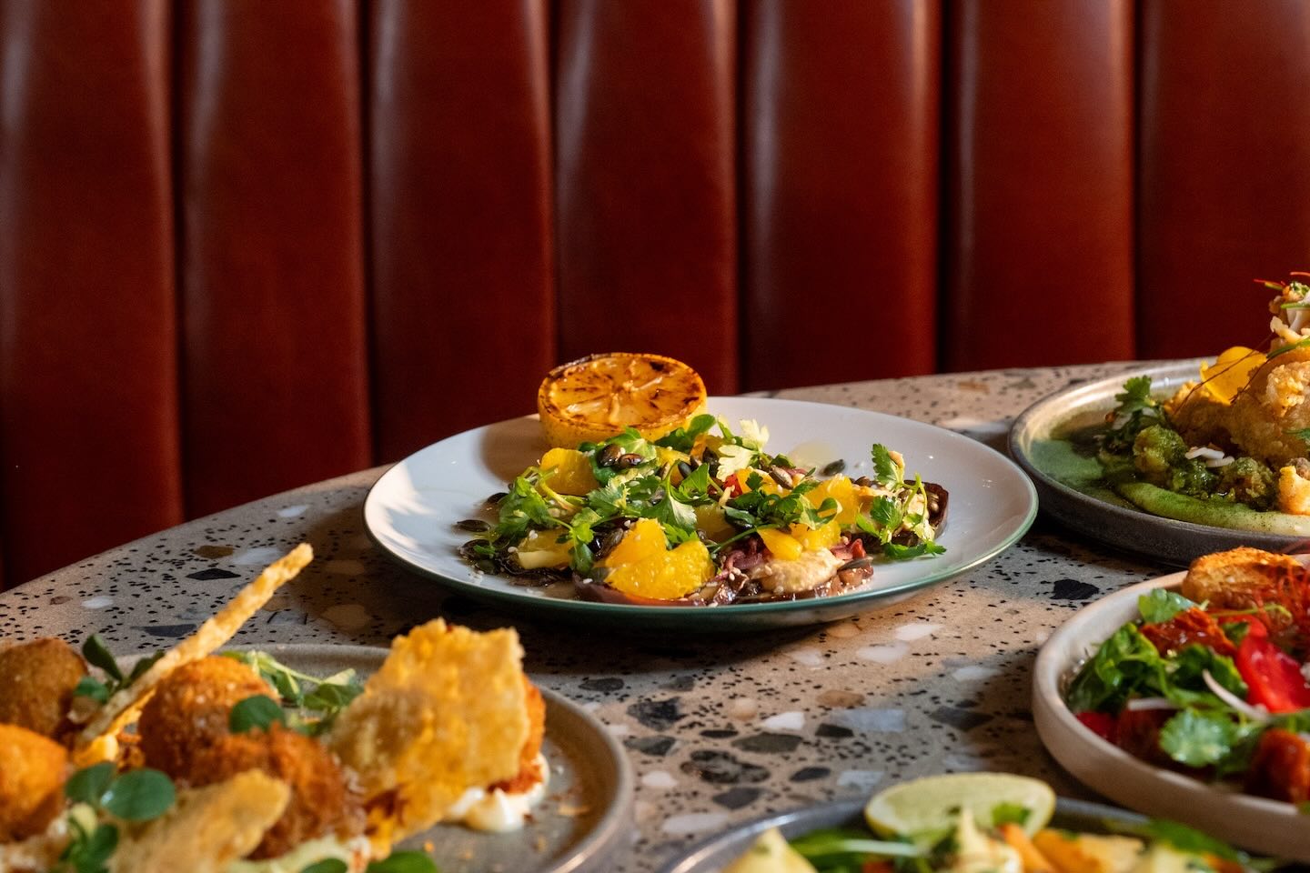 A shallow depth-of-field shot focusing on a salad of citrus segments, pumpkin seeds, and fresh greens. A charred lemon half sits on the edge of the white plate.