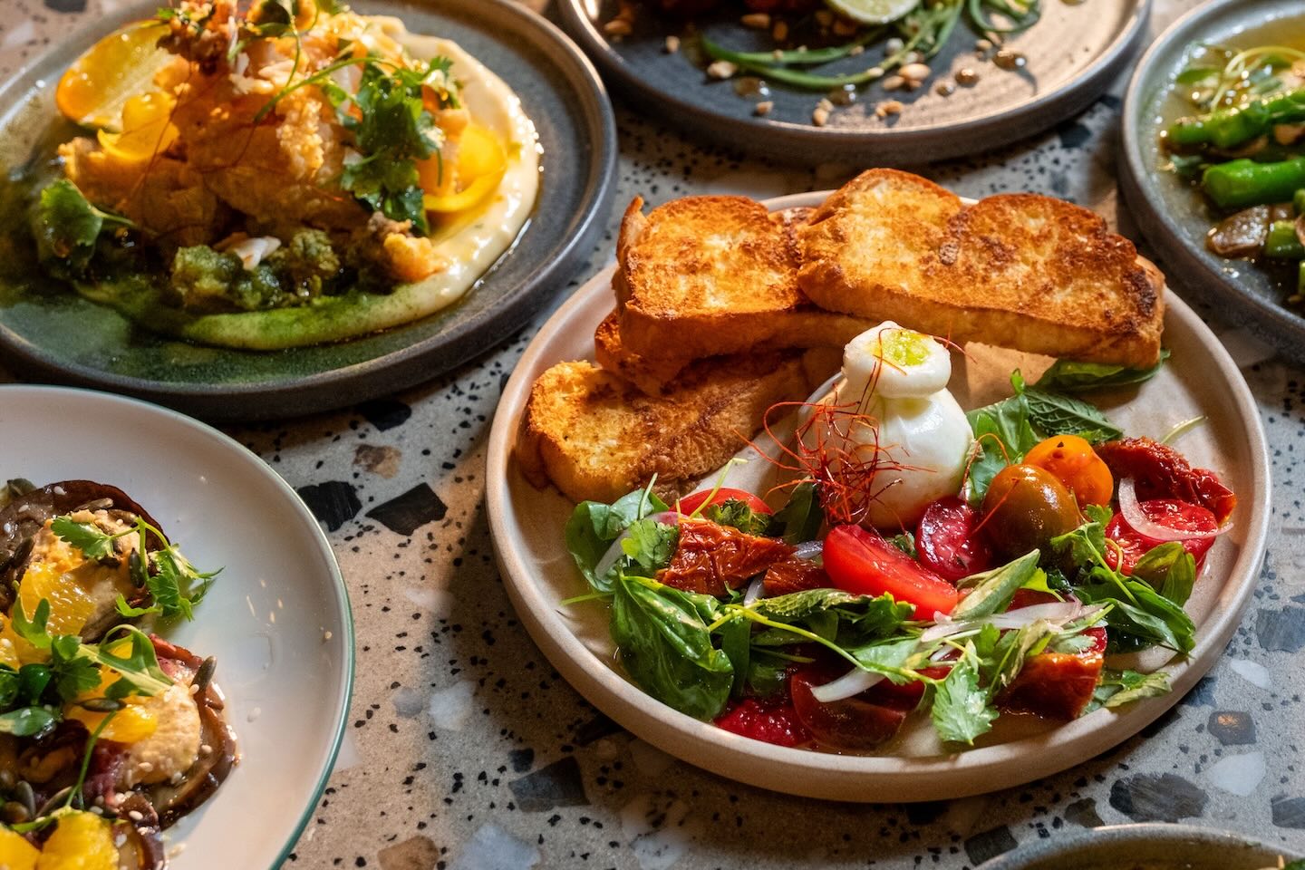 A close-up of a burrata salad featuring vibrant red and yellow tomatoes, fresh basil, and balsamic-marinated onions, served with three thick slices of golden-brown toasted brioche.