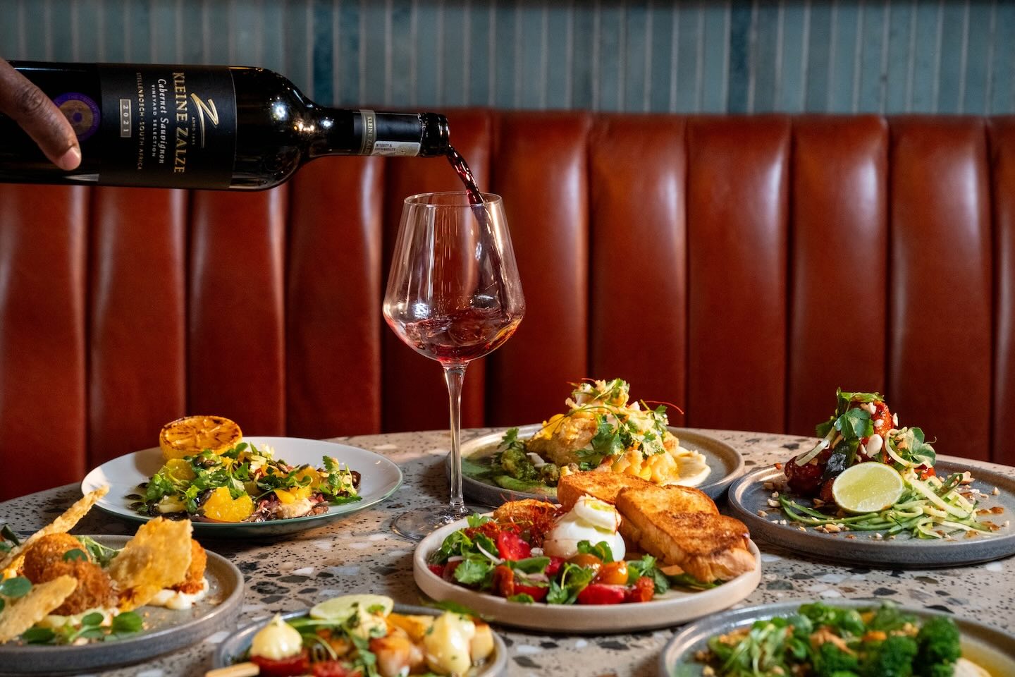 A person pours red wine from a bottle of Kleine Zalze Cabernet Sauvignon into a tall glass. The table is filled with colorful Mediterranean-style small plates, set against a background of a tufted cognac leather banquet.