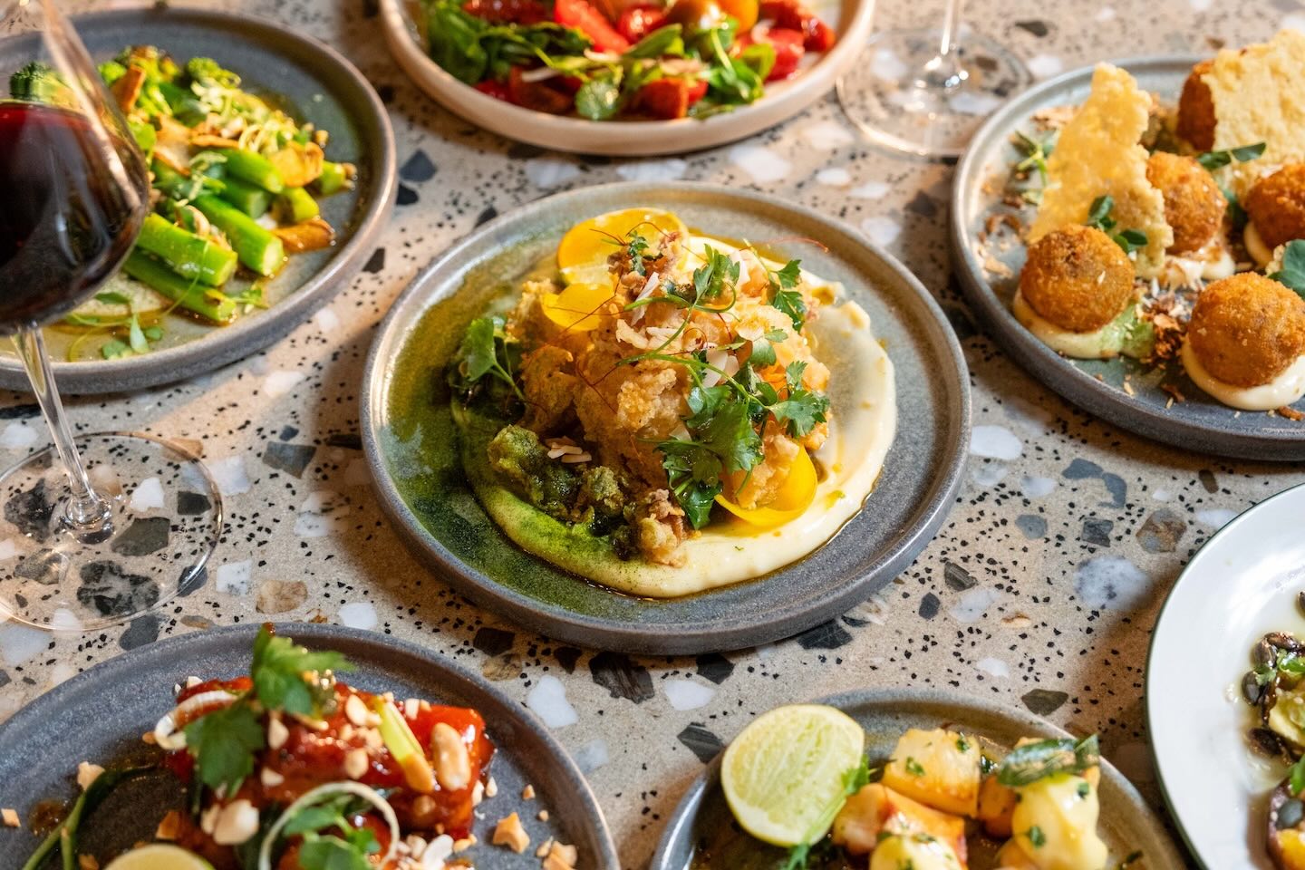 A high-angle view of a stone table crowded with appetizers. The central focus is a mound of crispy fried seafood garnished with fresh herbs and citrus, surrounded by dishes of grilled vegetables, skewers, and arancini.