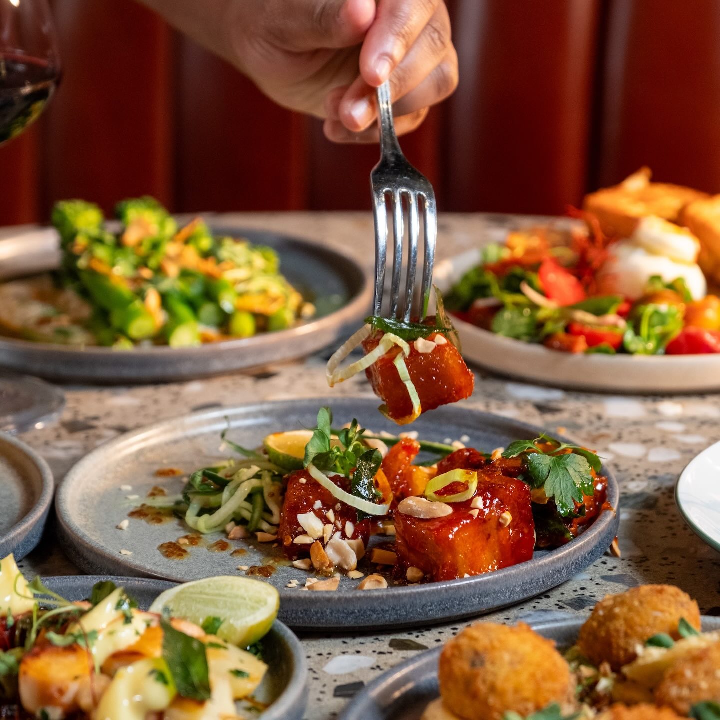 A close-up of a hand using a silver fork to lift a single cube of glazed, deep-red pork belly from a grey ceramic plate. The dish is garnished with crushed peanuts, sliced scallions, and a side of pickled cucumber ribbons.