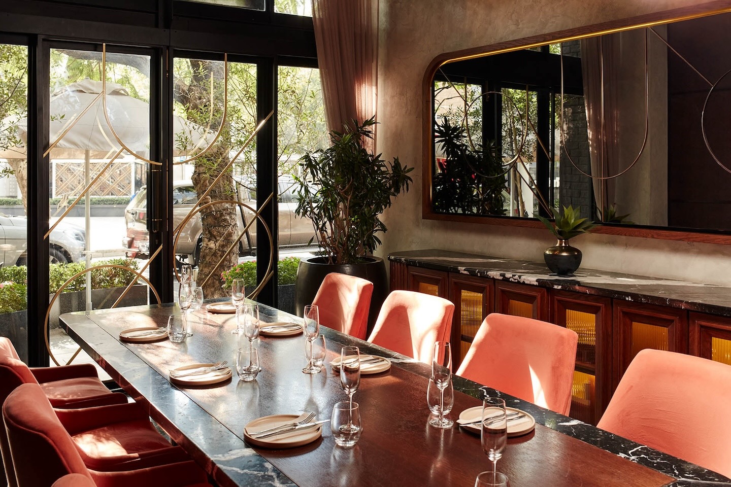 A sun-drenched private dining room featuring a long dark wood table set for a formal meal with wine glasses and white plates. Plush coral-colored velvet chairs line the table, set against a large black-framed window and a statement mirror with gold geometric overlays