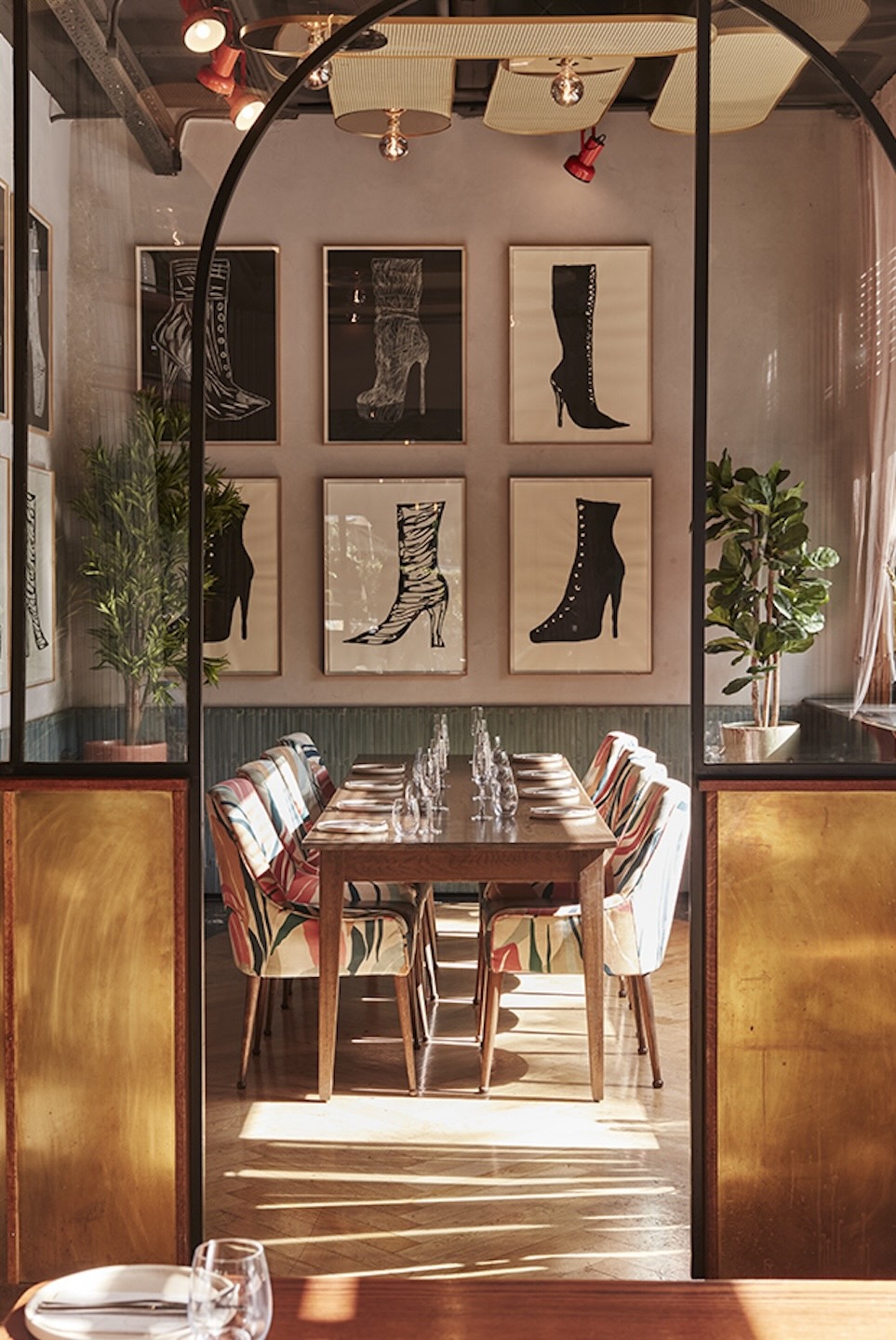 A bright, boutique private dining space viewed through a gold-accented glass partition. The room features chairs with a colorful tropical leaf pattern and a gallery wall of six framed black-and-white illustrations of high-heeled boots.