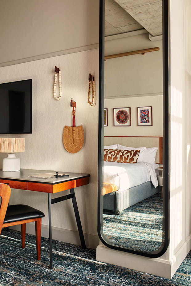 Interior view of a hotel room featuring a mid-century modern wooden desk with a black leather chair. A full-length black-framed floor mirror leans against the wall, reflecting a king-sized bed with patterned pillows and framed African-inspired artwork. Woven decorative items hang on the neutral-toned wall.