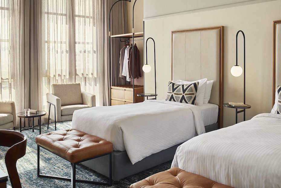 A modern VIP twin hotel room featuring two single beds with white striped linens, tan leather tufted benches at the foot, and sleek arched black floor lamps. Large windows with sheer curtains fill the room with natural light.