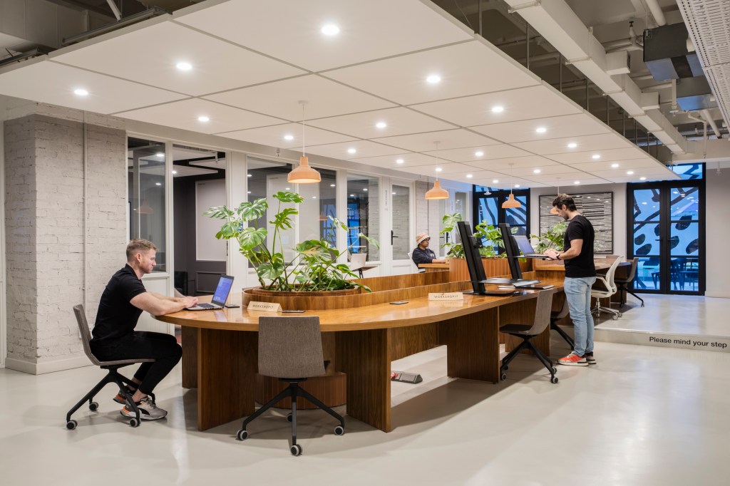 A bright, open-plan office featuring a long, curved wooden desk with built-in leafy green planters. Professionals are working on laptops under a large rectangular ceiling light panel. Glass-walled private offices line the background.
