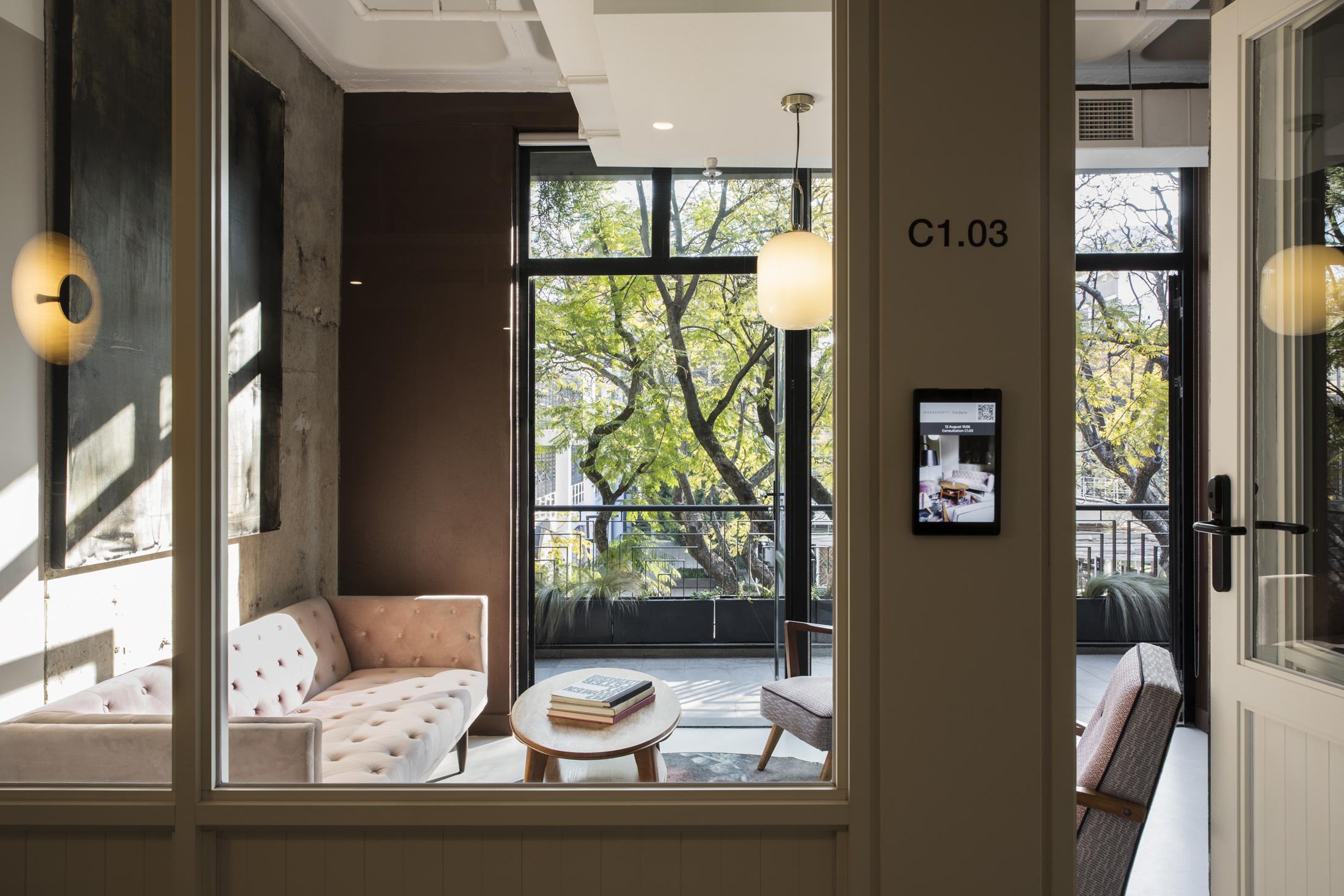 A view through a glass door into a cozy private meeting room labeled C1.03. The room contains a plush velvet sofa, a small coffee table, and large windows looking out onto lush green trees.