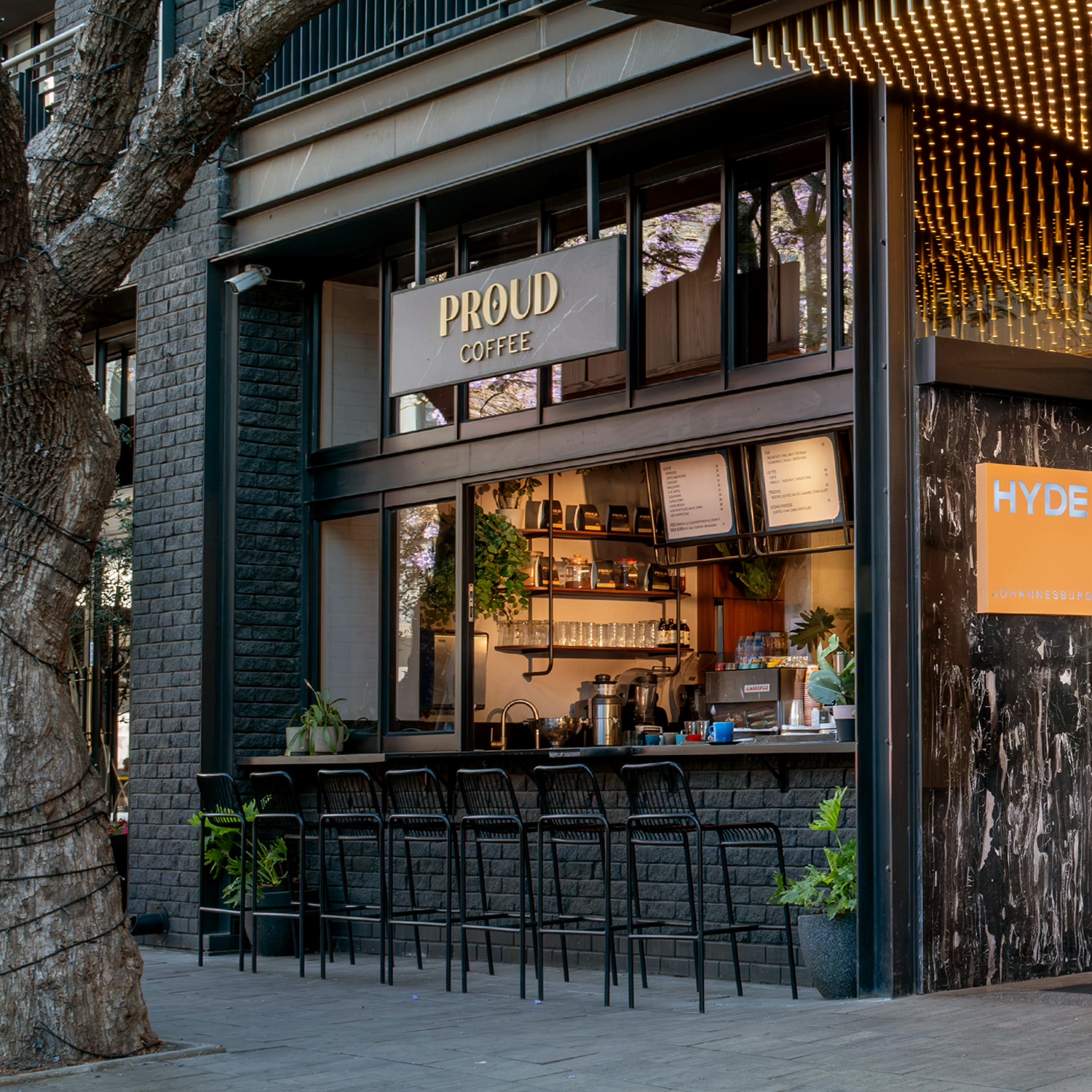 An outdoor view of "Proud Coffee" featuring a black brick walk-up counter with barstools.