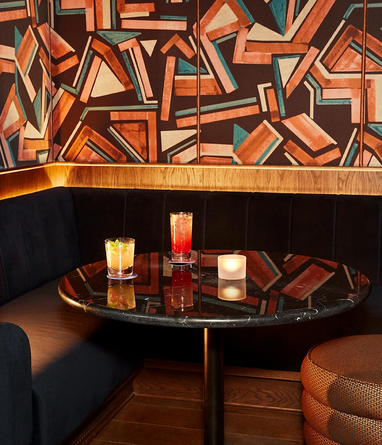 Close-up of a circular black marble table in a dimly lit bar booth. Two colorful cocktails and a frosted votive candle sit on the table, reflecting off the polished surface. The background features a dark velvet banquette and a wall covered in abstract, geometric wallpaper in shades of burnt orange, teal, and brown.