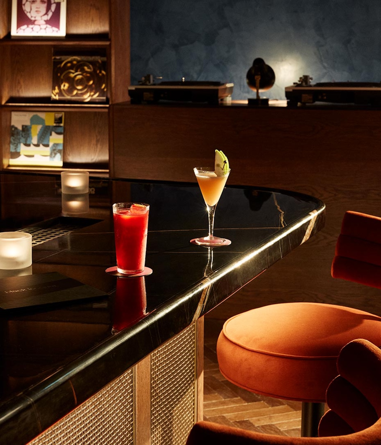 A high-angle shot of a sleek black marble bar top featuring a bright red cocktail and a light-colored martini garnished with an apple slice. In the background, a wooden shelf holds vinyl records, and two turntables are visible on a DJ booth against a textured blue wall.