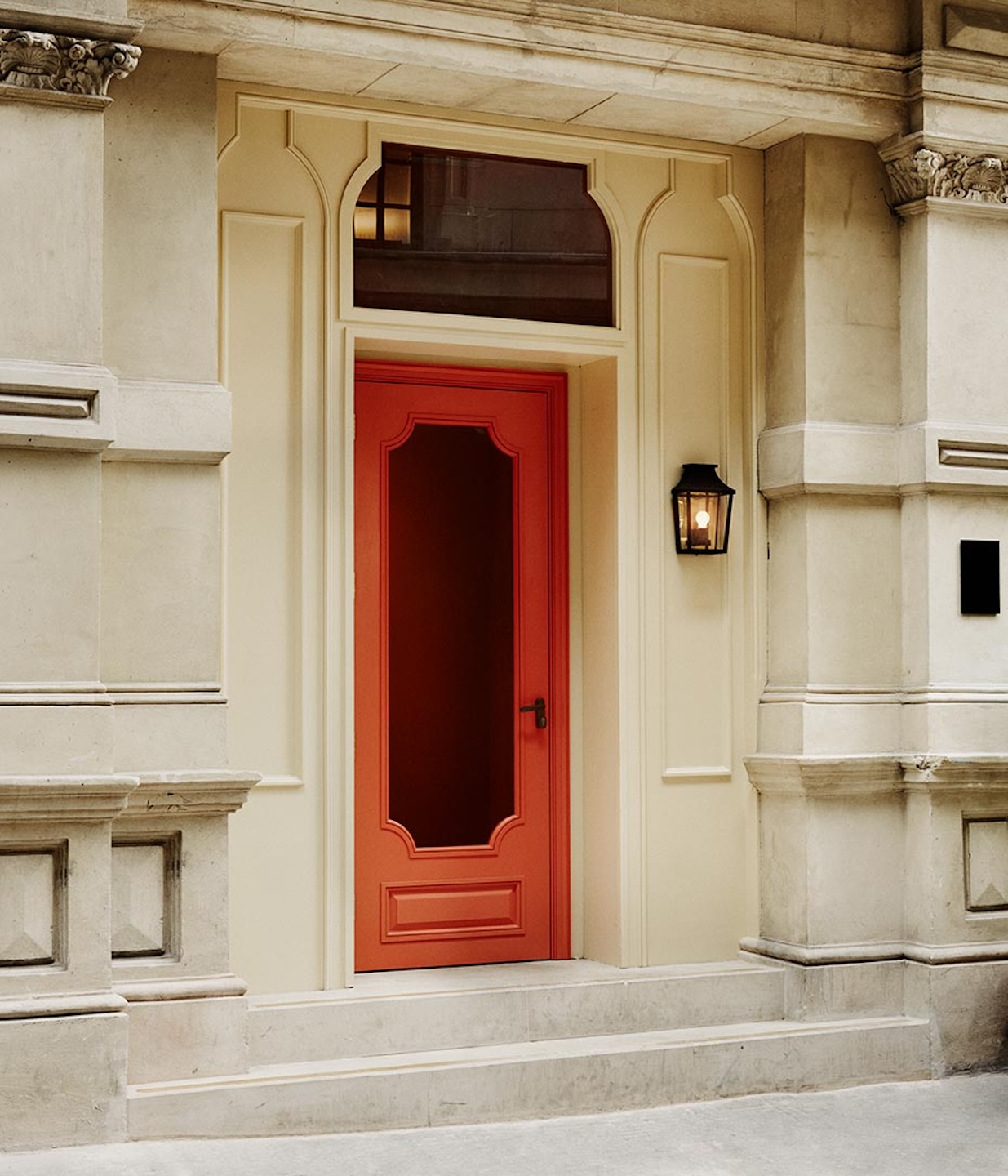A vibrant coral-red door with a tall, narrow glass pane set into a classic cream-colored stone facade. The entrance is flanked by ornate architectural pillars and illuminated by a traditional black lantern mounted on the wall.