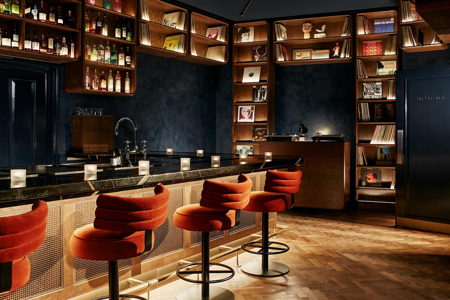 A wide view of a sophisticated cocktail bar featuring plush, red velvet stools lined up against a black marble counter. Warm wooden shelving filled with vinyl records and spirits wraps around the dark blue textured walls, illuminated by small glowing candles.