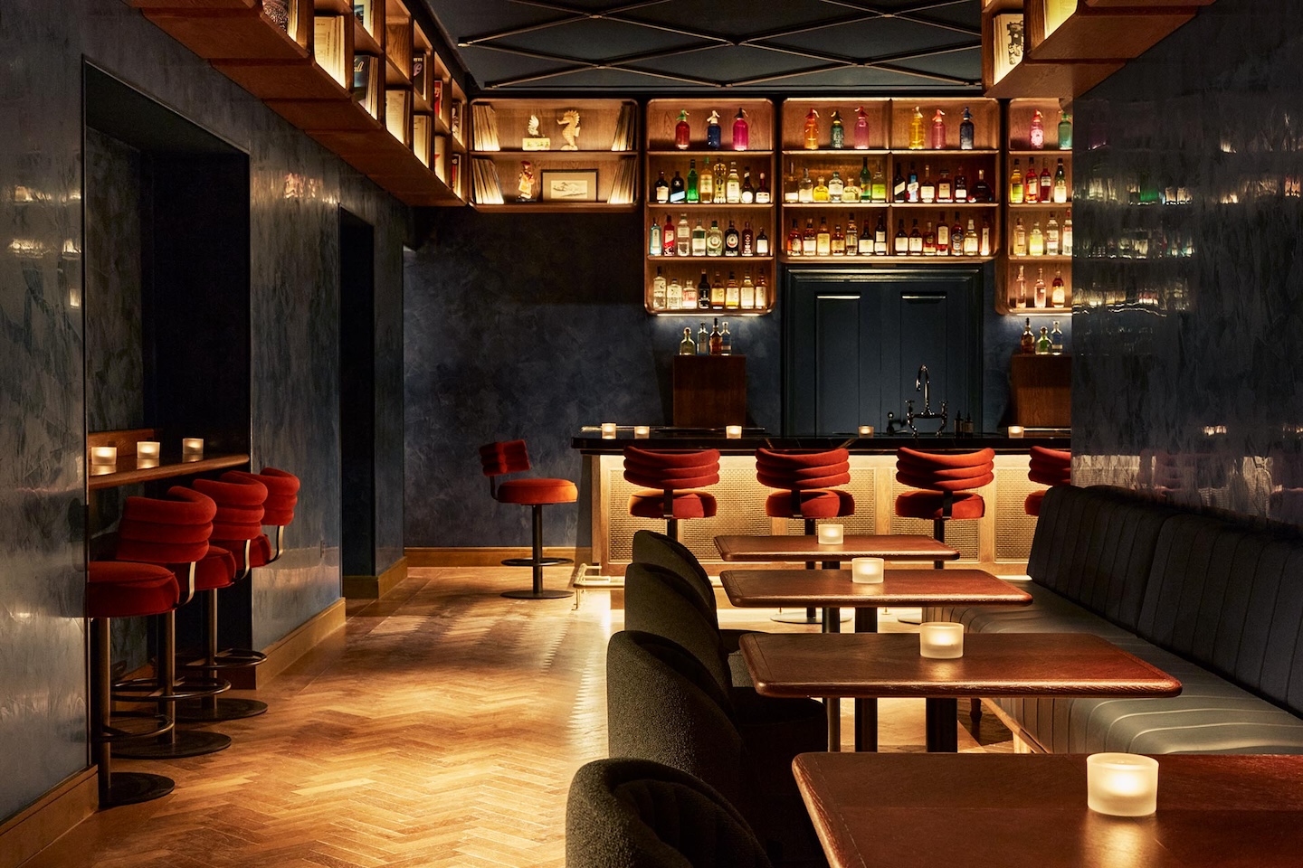 The interior of a moody, upscale lounge featuring herringbone wood floors and dark blue walls. The space includes a bar area with red stools, a separate seating area with small wooden tables and a long banquette, and shelves lined with vinyl records and colorful bottles.