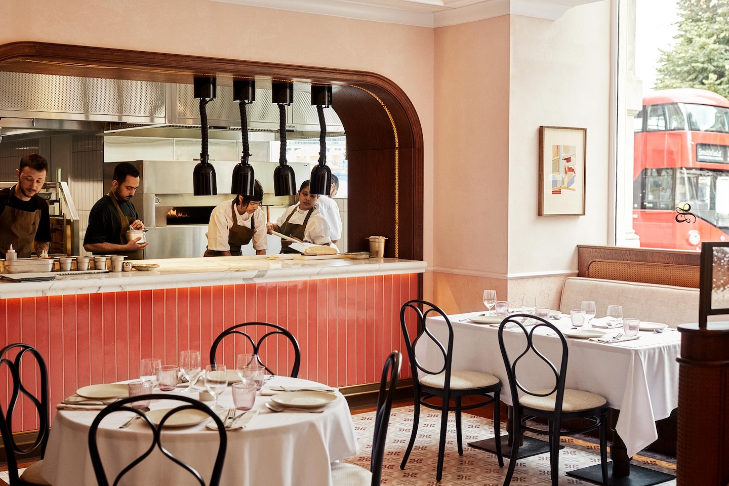 An open kitchen view from a dining room with white-clothed tables and black bistro chairs. Chefs are visible through a large arched pass with a red-tiled base. In the background window, a classic red double-decker London bus is partially visible.