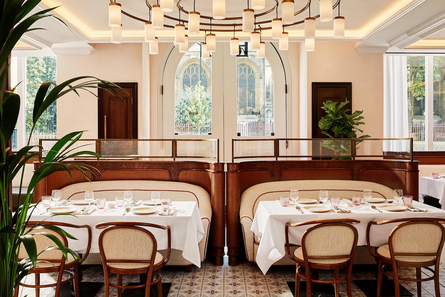 Symmetrical view of two dining booths with curved wooden frames and cream upholstery. The tables are set with white linens, glassware, and pink water glasses. Large arched windows in the background reveal a view of a stone church building.