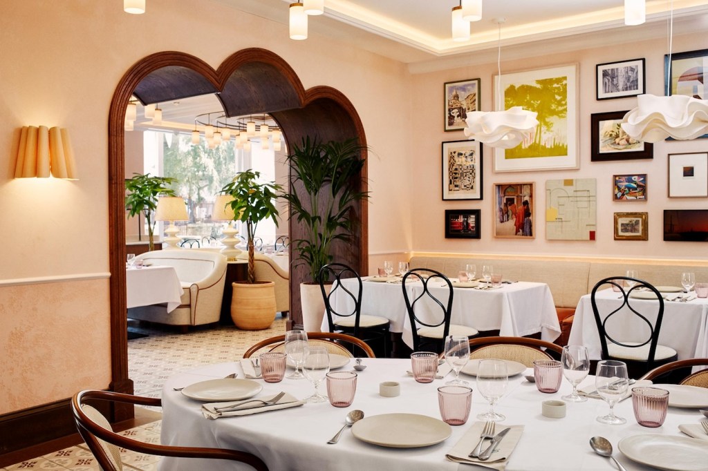 A spacious dining room with various seating arrangements, including round and rectangular tables with white tablecloths. The walls are decorated with a gallery of framed art and unique wavy white pendant lights. A decorative wooden archway leads into another section of the restaurant.