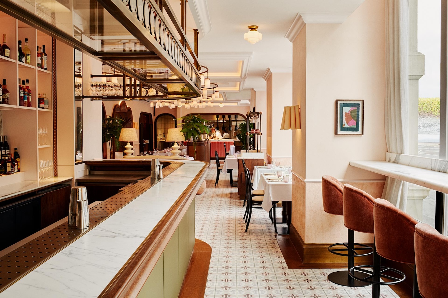 A perspective shot looking down the length of the bar toward the dining area. The white marble bar top is in the foreground, with velvet salmon-colored bar stools positioned along a window ledge to the right. The patterned tile floor leads the eye toward the warm, lit interior.