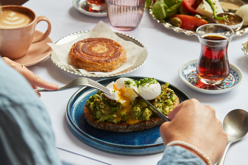 A close-up, top-down view of a breakfast table featuring a person cutting into a poached egg on avocado toast. The yolk is runny, and the toast sits on a dark blue plate. Surrounding the main dish are a latte in a pink cup, a small glass of dark tea, a pastry on a silver plate, and a platter of fresh vegetables and cheese.