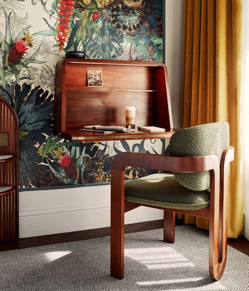 A green desk chair with dark wooden arms and legs, pulled out from a dark wooden desk with sunlight hitting it's back from the window beside