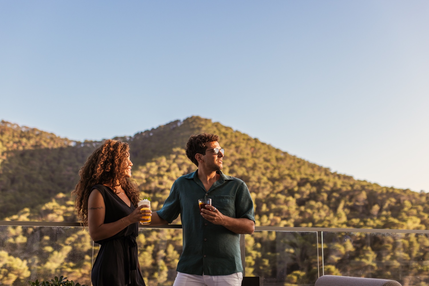 two people standing by the rail of the terrace holding a drink in their hand with a cliff in the background
