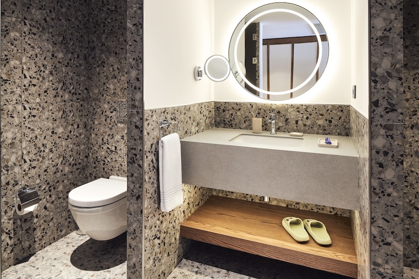 Grey bathroom sink with circular mirror and wooden shelf with sandals on it