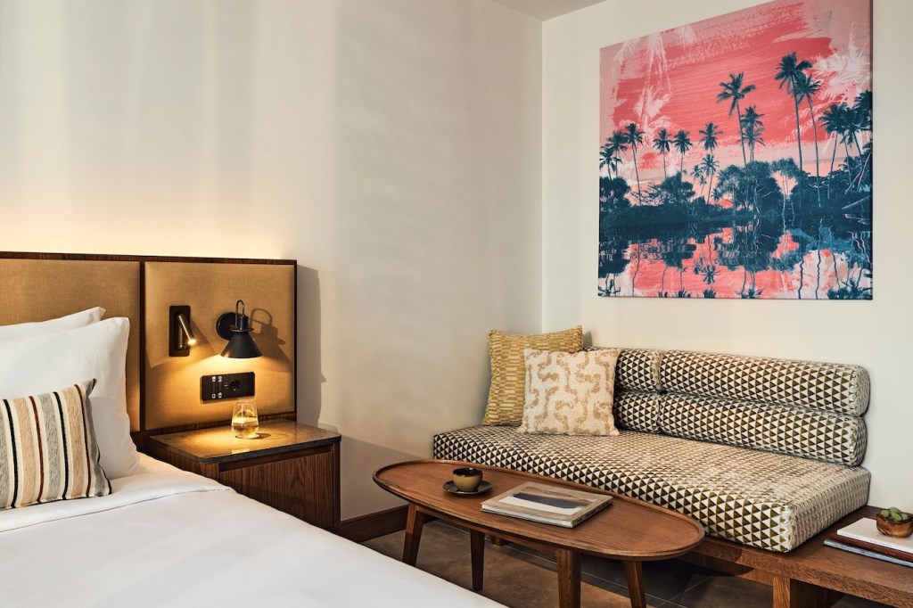 Angled view of large bed with beige headboard, bedside table, light, coffee table, and upholstered bench with colorful artwork on the wall