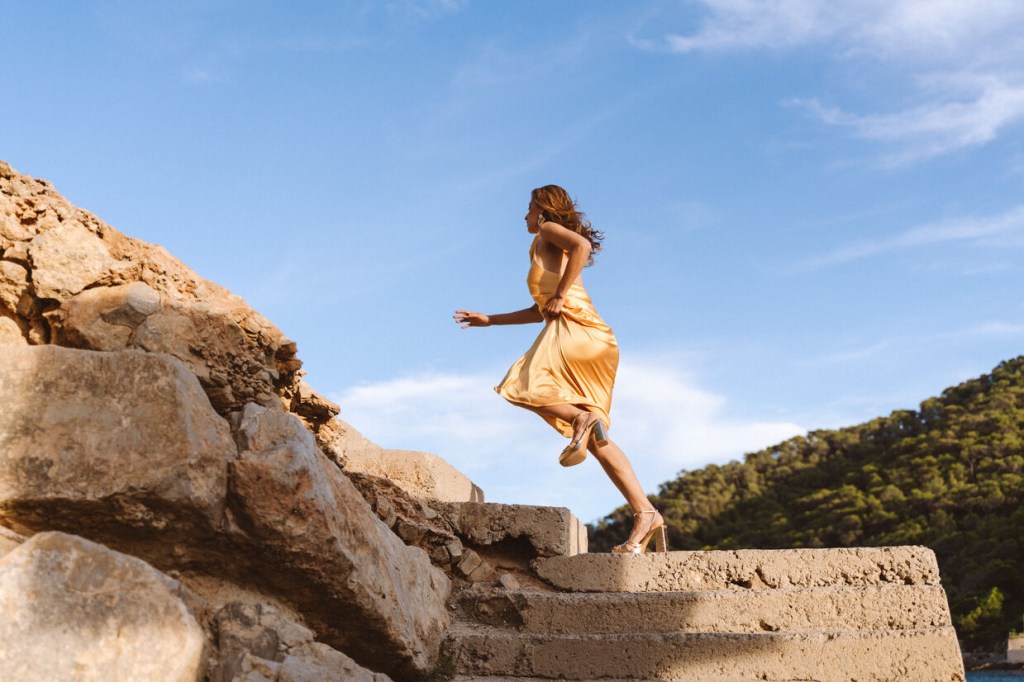 Young woman running upwards through stairs located at the Cala Llonga bay. She is wearing a gold dress.