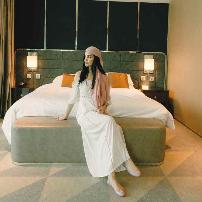 Woman in long white dress sits on a large bed with gray headboard