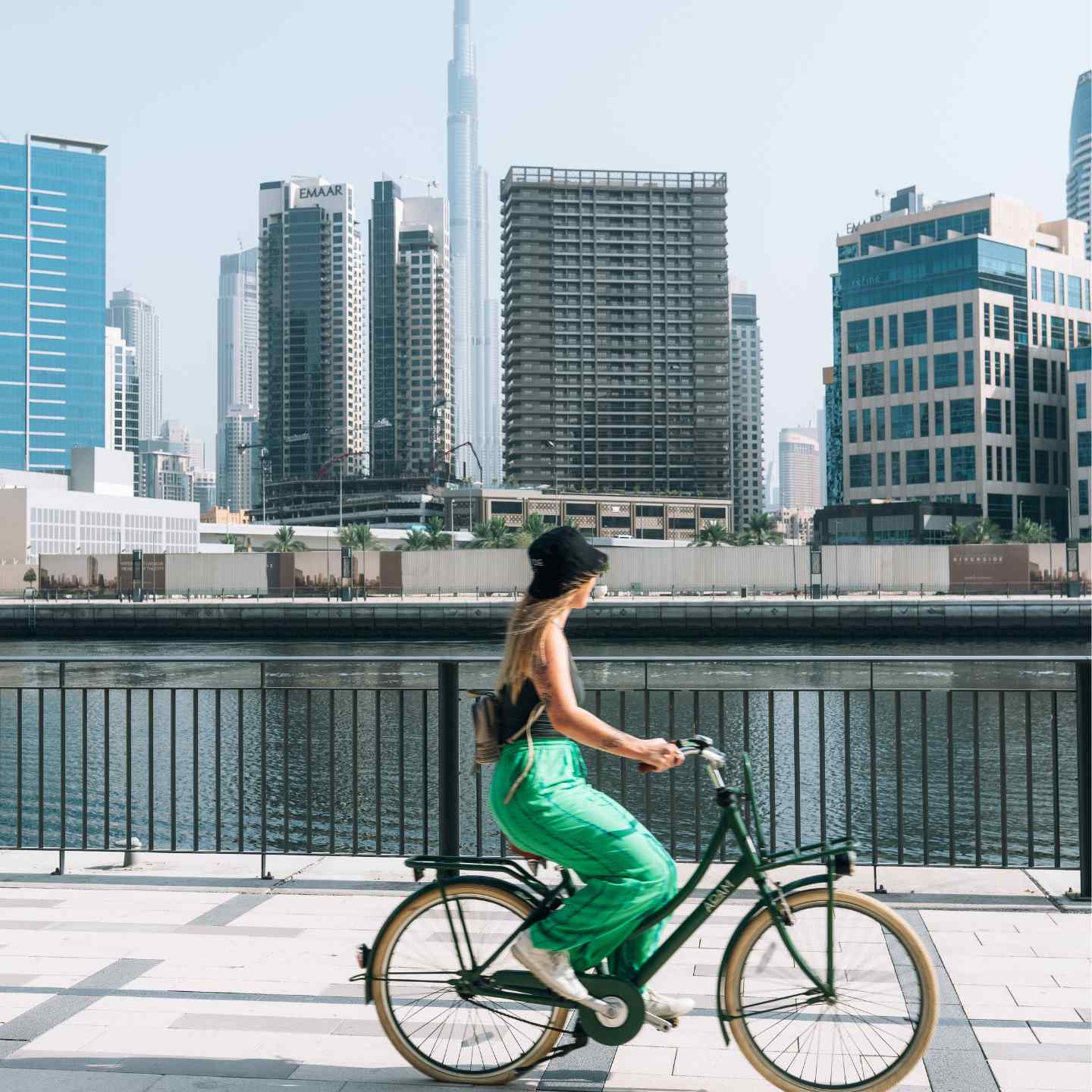 A woman in a bright green skirt riding a bicycle along the Dubai waterfront with a canal and high rise buildings in the background