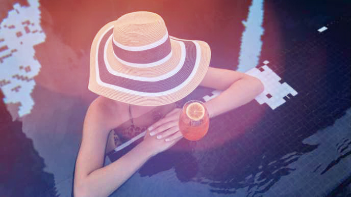 Woman in sunhat with arms on pool ledge and orange cocktail nearby